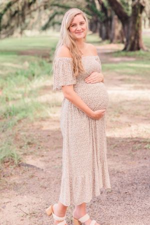 Expectant mother in flowy off-shoulder dress poses on natural pathway surrounded by oak trees.
