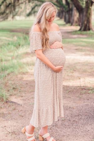 Mother-to-be in delicate floral dress cradles her bump while standing on a nature trail.