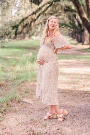Joyful maternity portrait of expectant mother laughing on tree-lined path in floral dress.