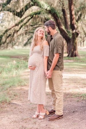 Romantic maternity moment between couple on oak-lined path during outdoor photo session.