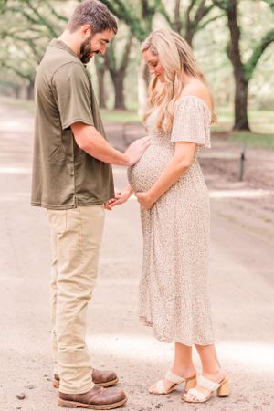 Sweet maternity moment captured on a tree-lined path as couple looks down at baby bump.