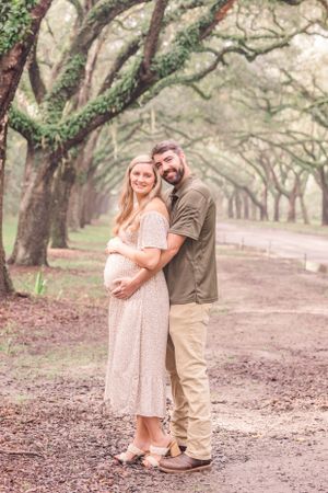 Expecting couple embraces on a tree-lined dirt path during a romantic maternity photo session.