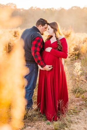 Romantic maternity photo of couple embracing in golden field at sunset wearing red buffalo plaid and dress.