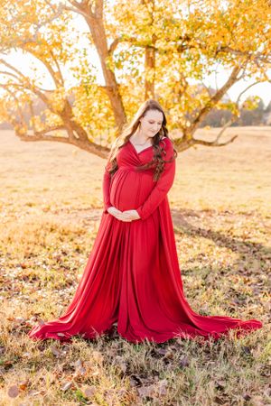 Maternity portrait in flowing red dress against autumn trees at sunset.