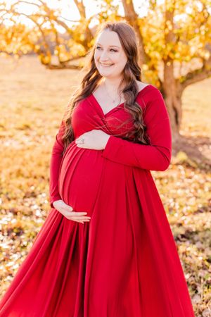 Woman in red maternity gown poses in golden autumn setting.