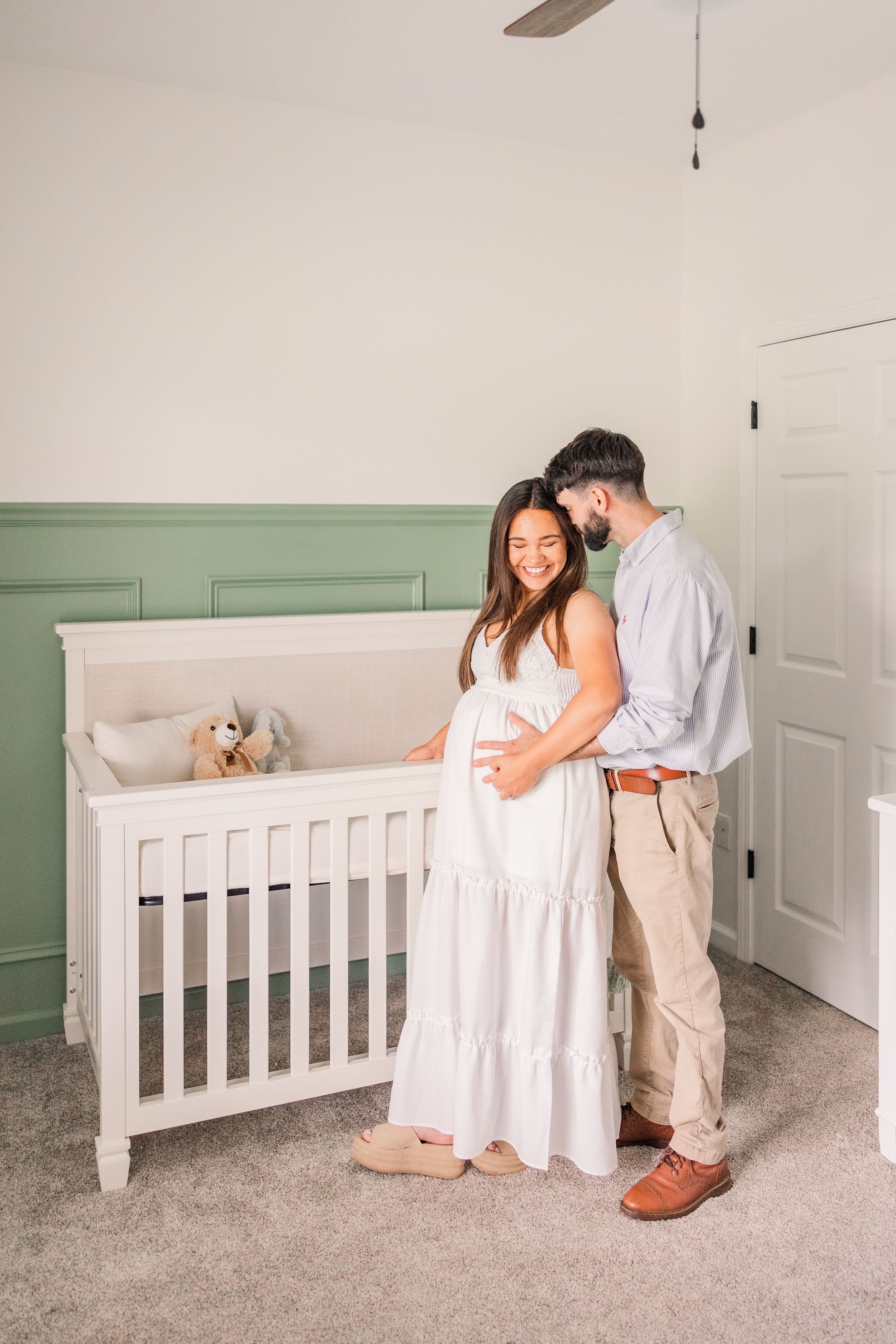 Expectant couple standing together in a nursery with a white crib and sage green walls.