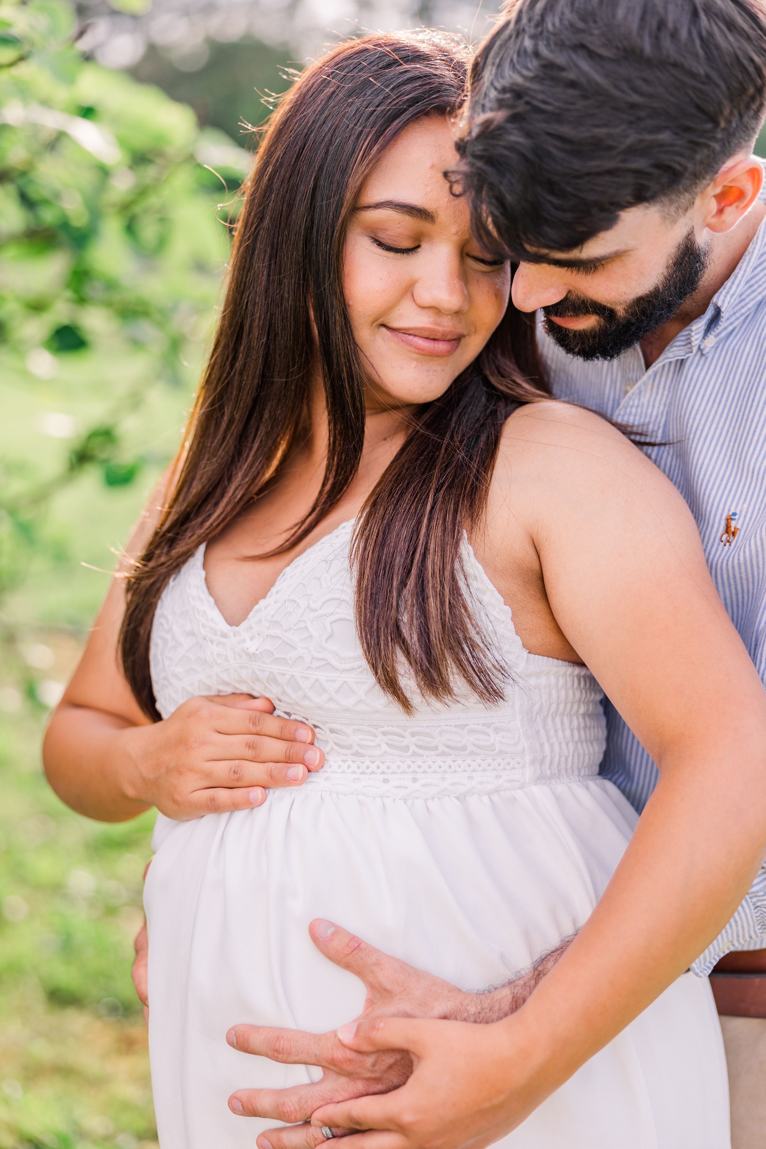 Intimate maternity portrait showing couple embracing while wearing white dress and blue shirt outdoors.