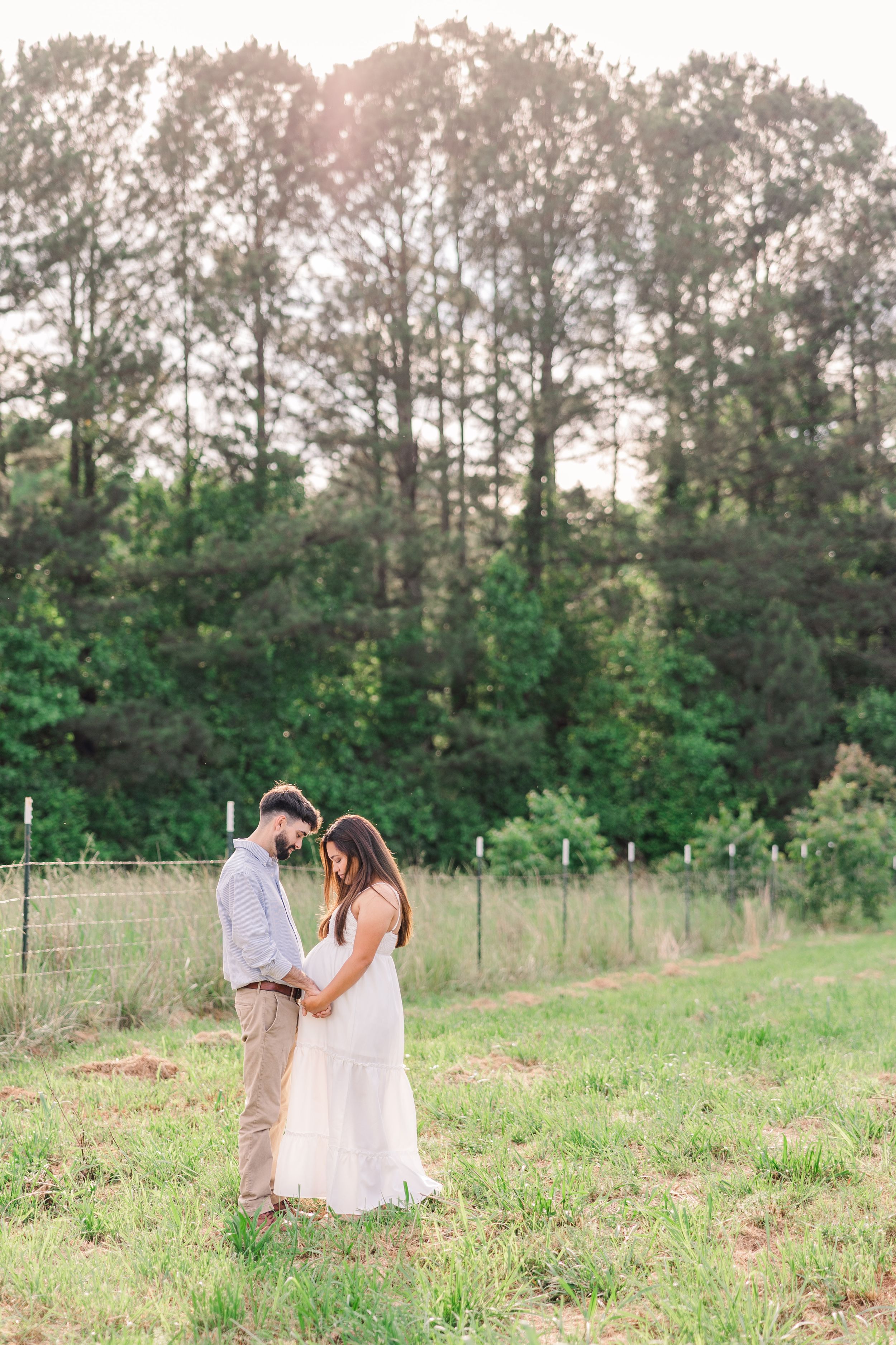 Couple shares tender moment in field with trees at sunset during maternity photoshoot.