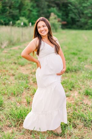 Maternity portrait of woman in flowing white dress standing in sunny outdoor field.