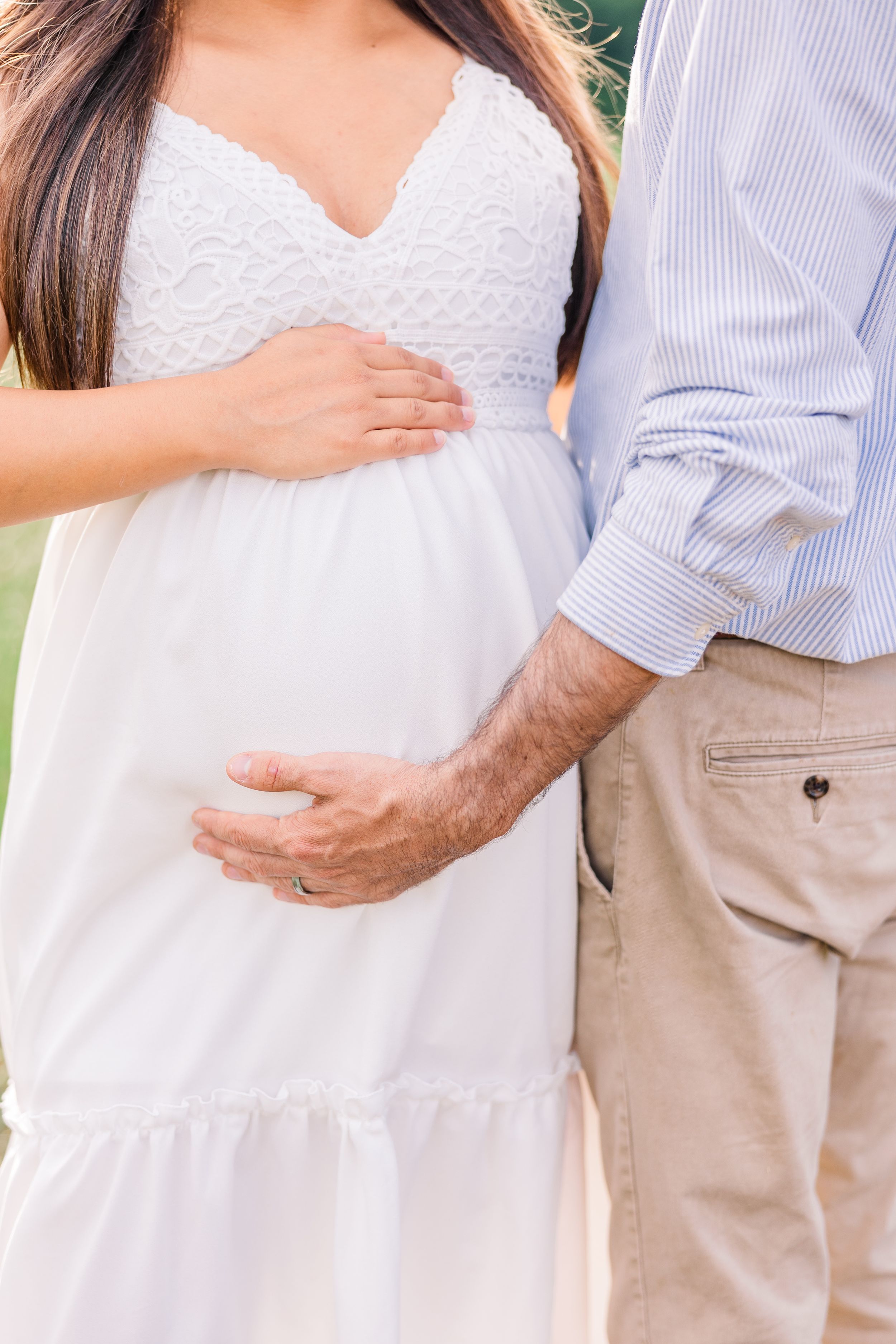 Close-up of couple's hands cradling a baby bump while wearing white dress and casual attire in outdoor setting.