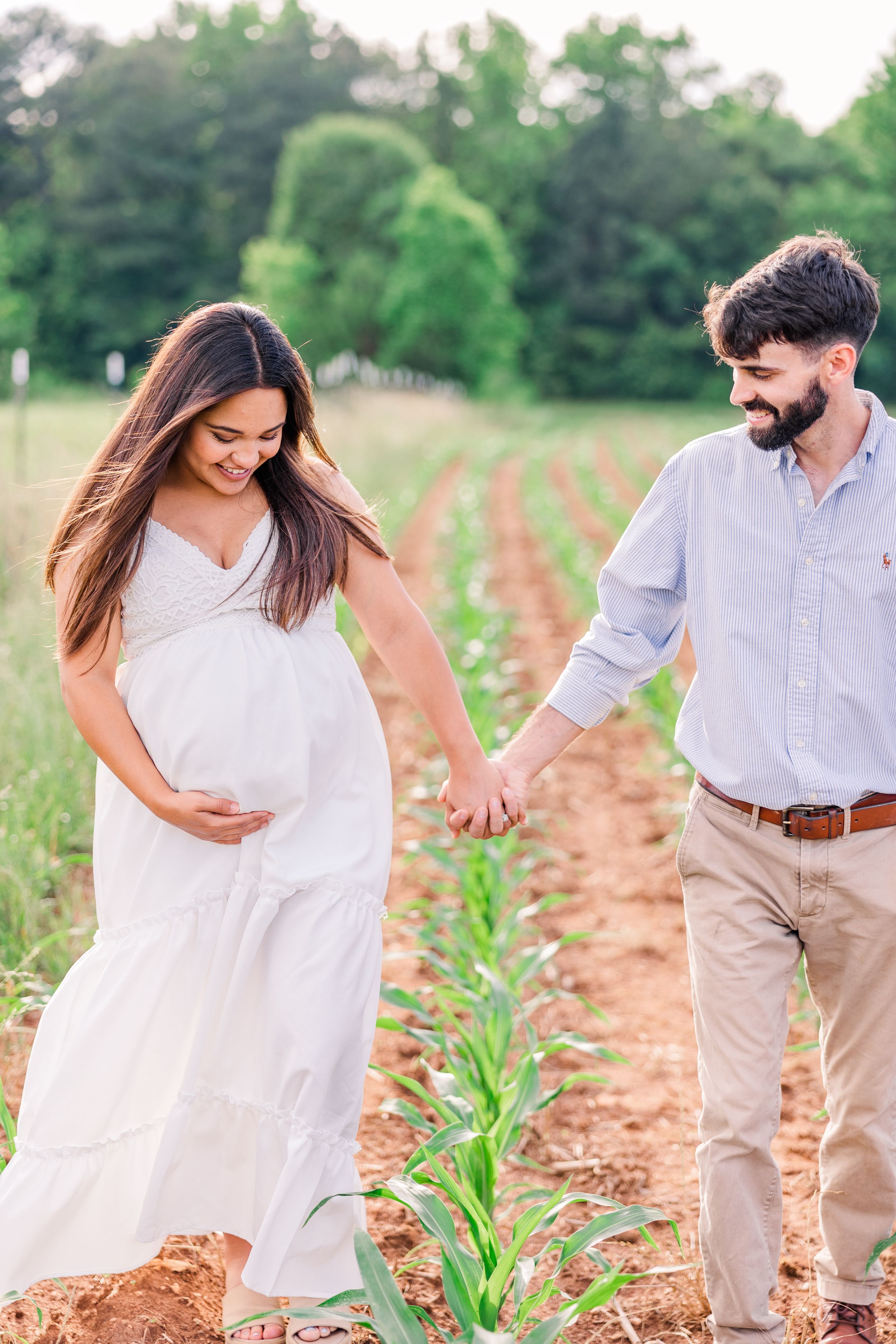 Expecting couple walks hand in hand through planted rows in a farm field.