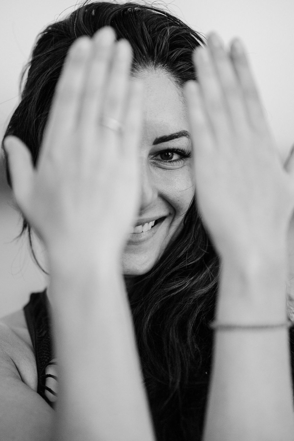 A playful black and white portrait showing hands covering half the face with a bright smile peeking through.