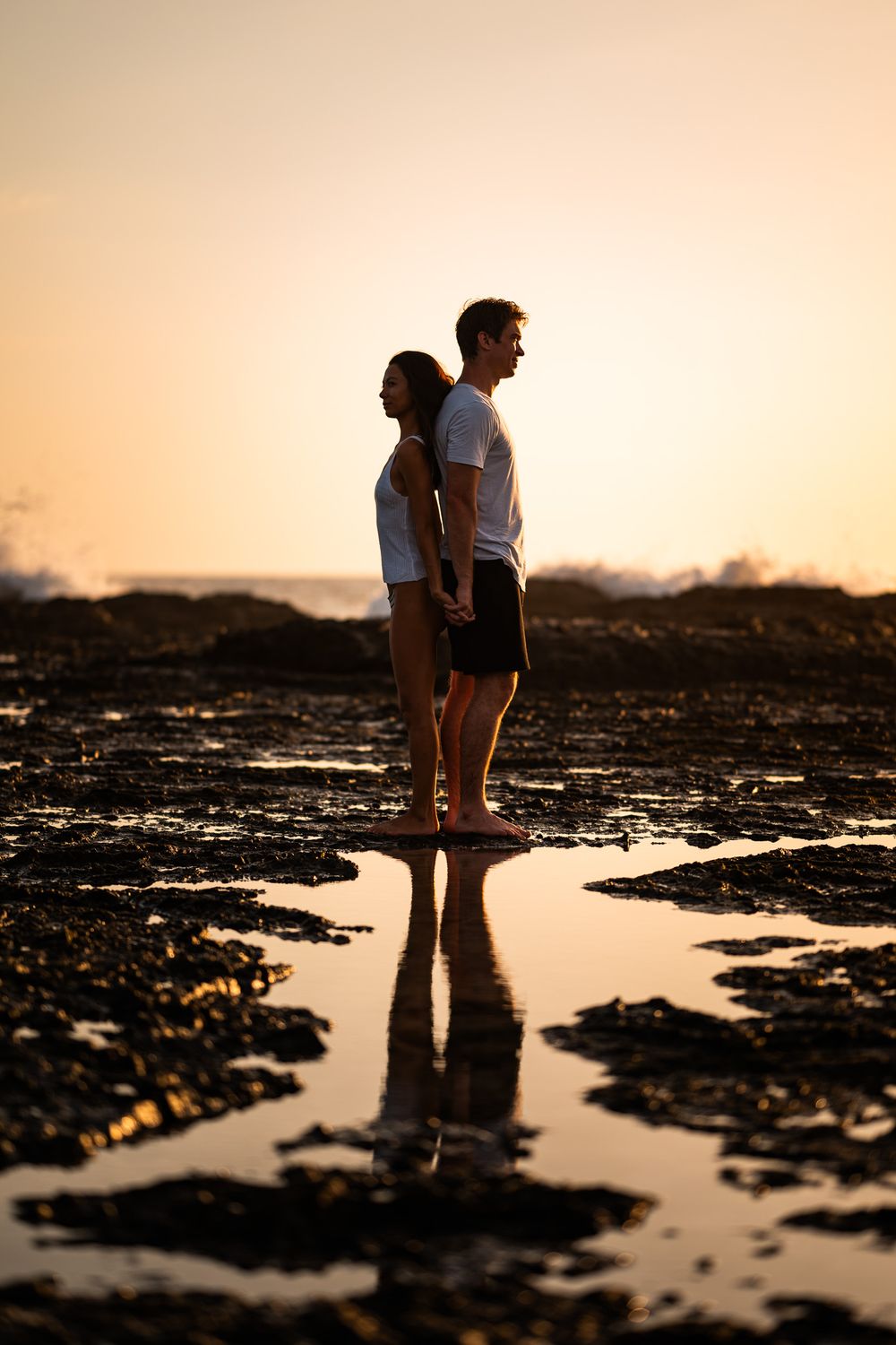 Couple silhouetted against golden sunset sky standing on rocky beach with reflection in tidepool.