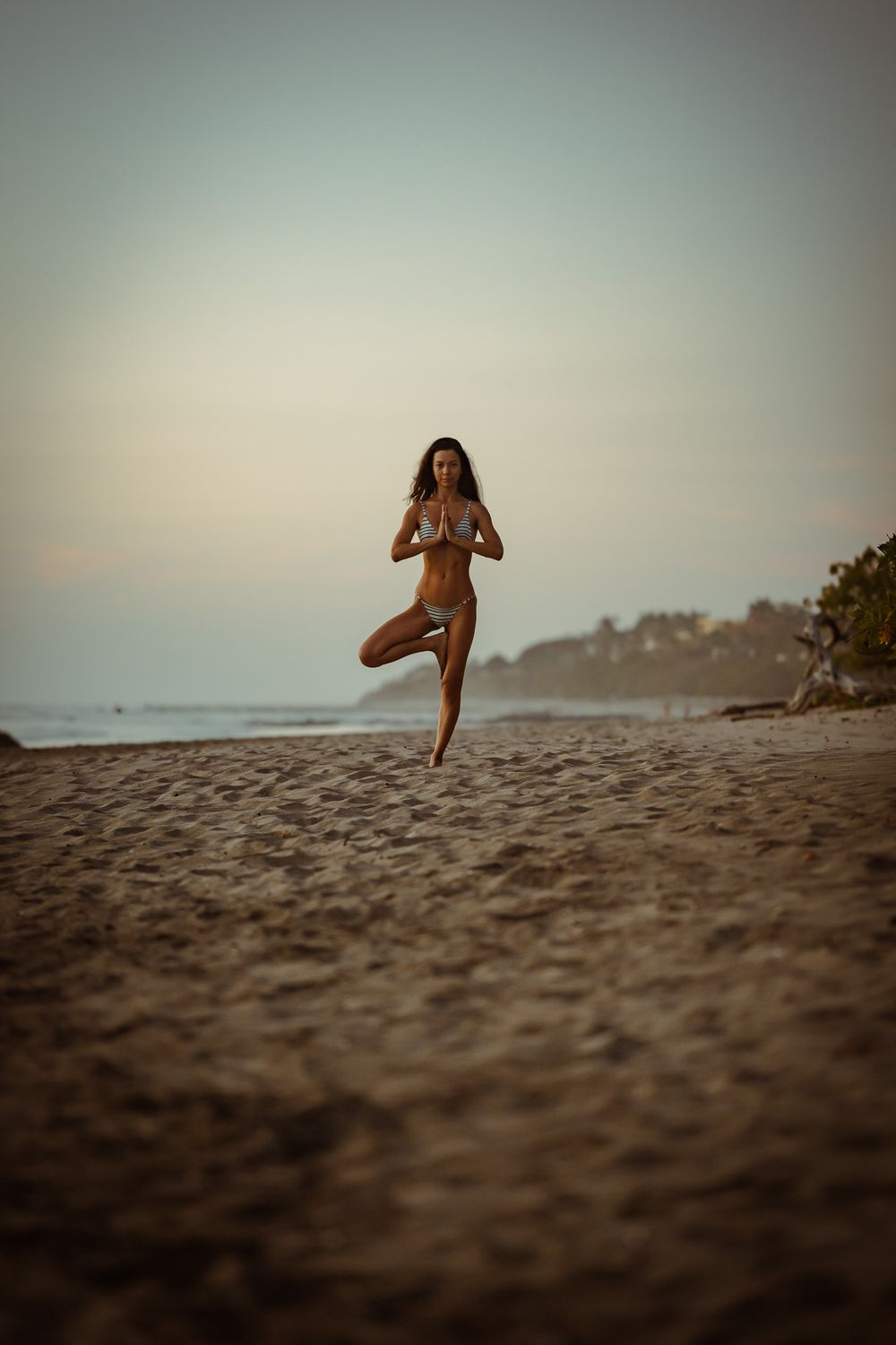 Woman practicing tree pose yoga on beach during sunset with coastal cliffs in background.