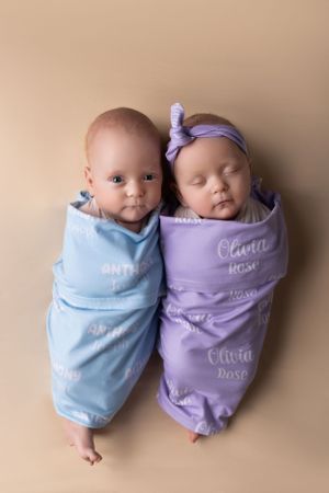 Two newborn infants swaddled in blue and purple blankets lying side by side on beige backdrop. NJ newborn photographer in Sussex County NJ