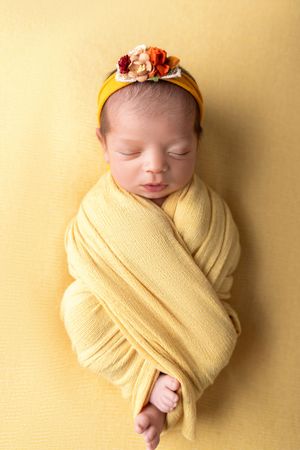 Sleeping newborn wrapped in soft yellow swaddle with floral headband against yellow background. NJ newborn photographer