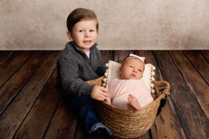 Two siblings pose for a portrait on wooden floor, with the younger one resting in a woven basket.