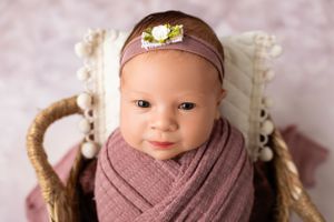 A newborn wrapped in a mauve swaddle blanket wearing a brown headband with yellow bow rests in a wicker basket.