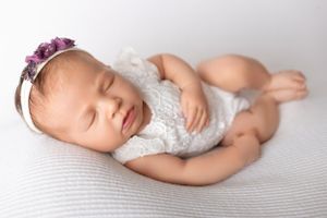 Newborn baby in white lace romper and floral headband sleeping peacefully on textured white blanket.