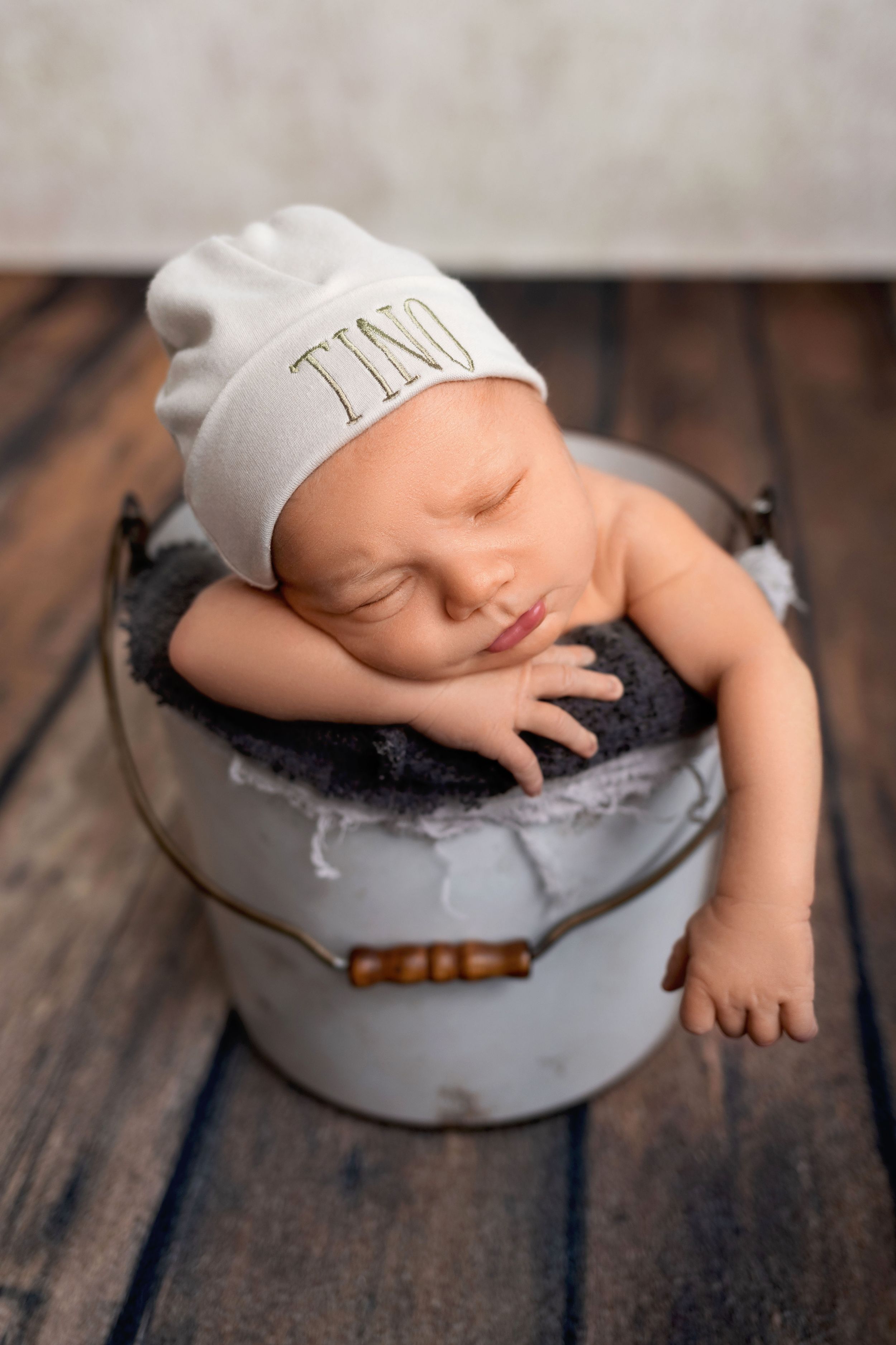 A sleeping baby in a white cap rests peacefully in a gray metal bowl on a rustic wooden surface.