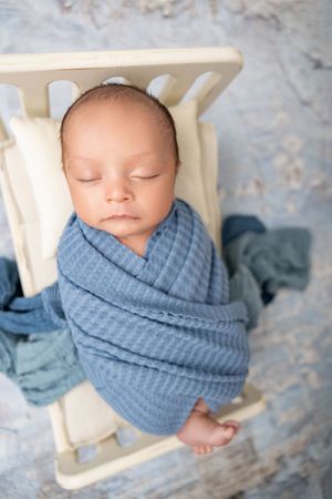 A sleeping newborn baby swaddled in soft blue knit fabric lies peacefully in a white wooden prop bed.