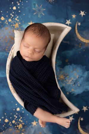 Newborn baby swaddled in navy knit wrap sleeps in white basket against starry night sky backdrop.