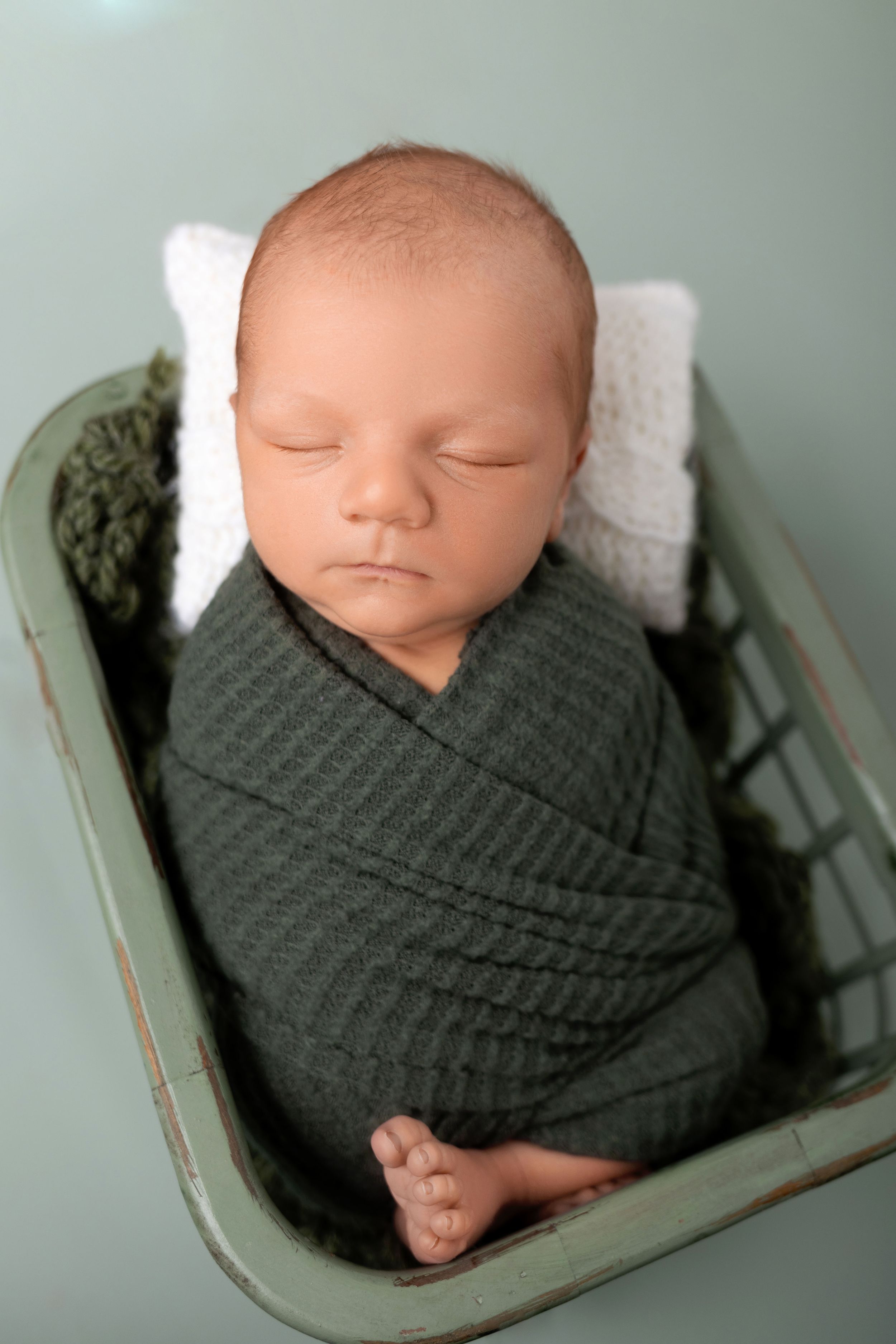A sleeping newborn swaddled in a dark gray knit blanket rests peacefully in a vintage mint-colored cradle.