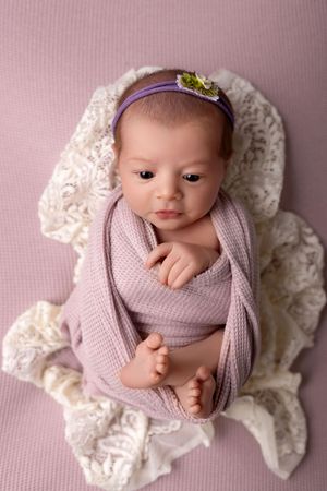Newborn baby swaddled in soft lavender fabric rests on white lace during professional portrait session.
