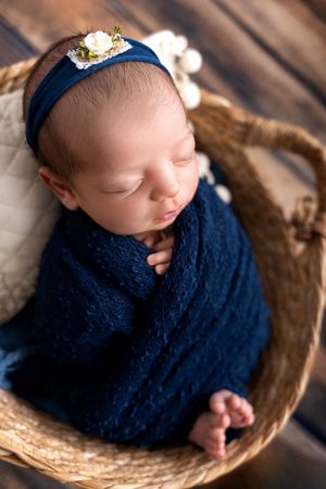Sleeping newborn wrapped in navy blue swaddle rests peacefully in woven basket wearing delicate headband.