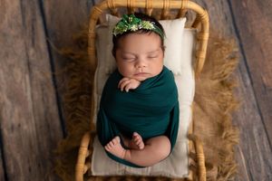 Sleeping newborn baby wearing dark teal wrap rests peacefully on wooden chair with rustic backdrop.