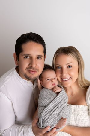 A loving family portrait with a newborn baby wrapped in gray swaddling between parents against a light background.