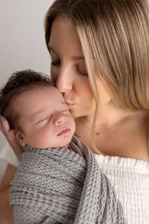 A tender moment as a mother kisses her sleeping newborn baby wrapped in a cozy gray knit blanket.