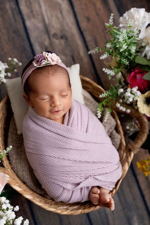 Sleeping newborn wrapped in soft purple swaddle rests in woven basket surrounded by delicate flowers on wooden floor.