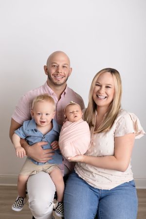 A family of four poses together against a light gray wall for a casual indoor portrait session.
