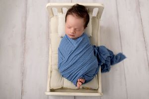 A sleeping newborn baby wrapped in soft blue fabric lies peacefully in a white wooden crate.