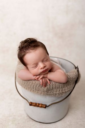 A sleeping newborn baby rests peacefully in a rustic white enamel bowl with a knitted beige blanket.