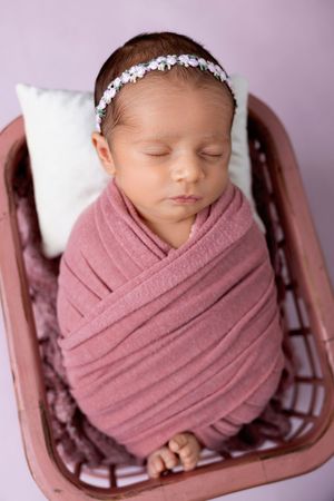 A sleeping newborn baby wrapped in dusty rose fabric and wearing a delicate headband rests in a wooden prop basket.