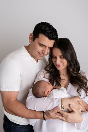 Parents in white clothing cuddle their newborn baby in a series of tender studio portraits.