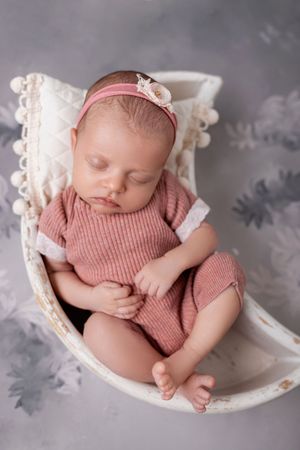 A sleeping newborn baby wearing a pink knit outfit rests peacefully in a white decorative bowl against a gray backdrop.
