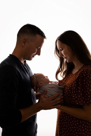 A silhouetted couple gazes lovingly at their newborn baby against a bright white background.