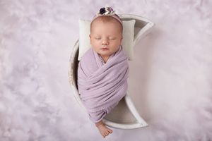 A sleeping newborn wrapped in soft lavender fabric lies peacefully in a white crescent-shaped prop against a purple backdrop.