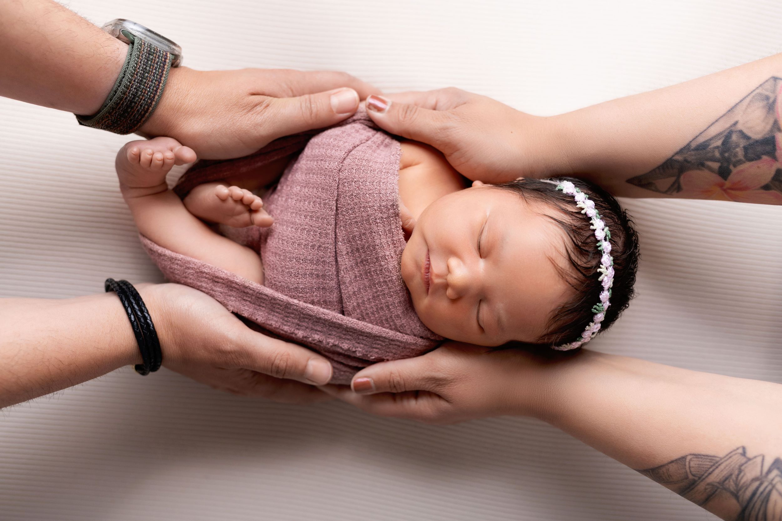 A series of hands gently holding and supporting a sleeping baby wearing a mauve knit outfit and delicate headband.