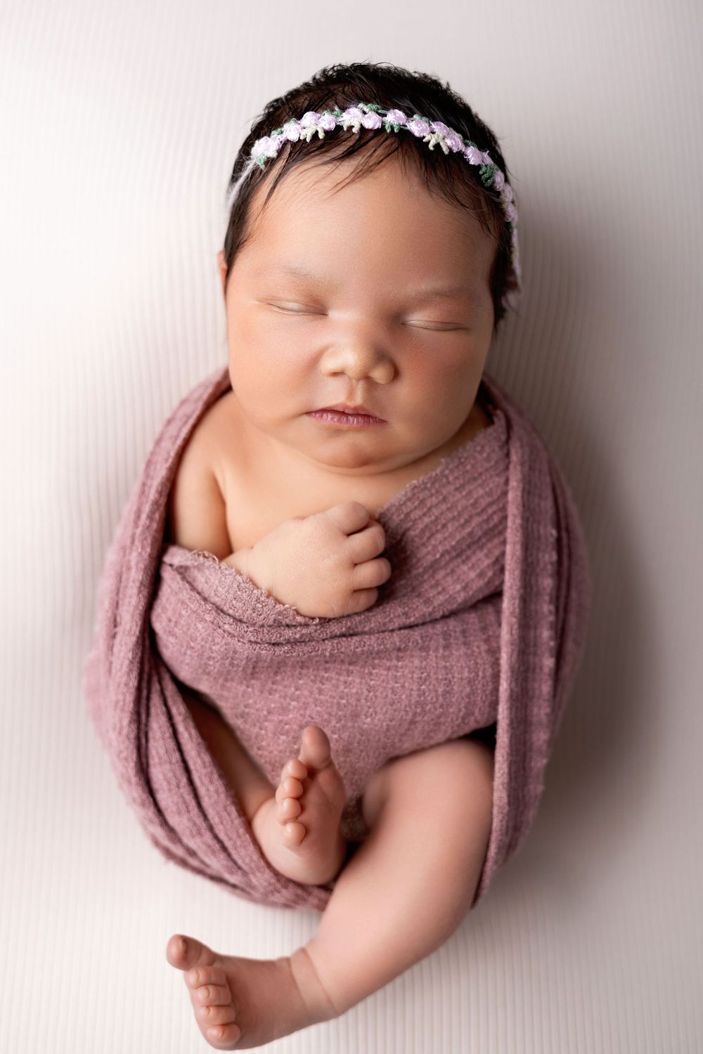 Newborn baby wrapped in dusty rose fabric lying on white backdrop with delicate floral headband in professional photo series.