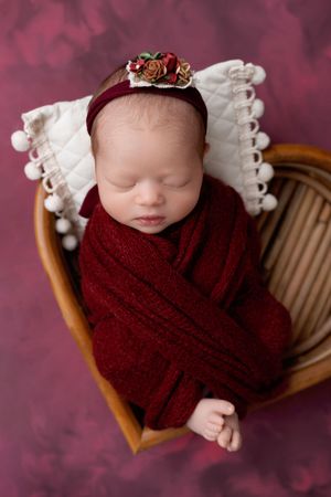 Newborn baby wrapped in burgundy blanket wearing white lace headband and floral crown resting in wooden chair.