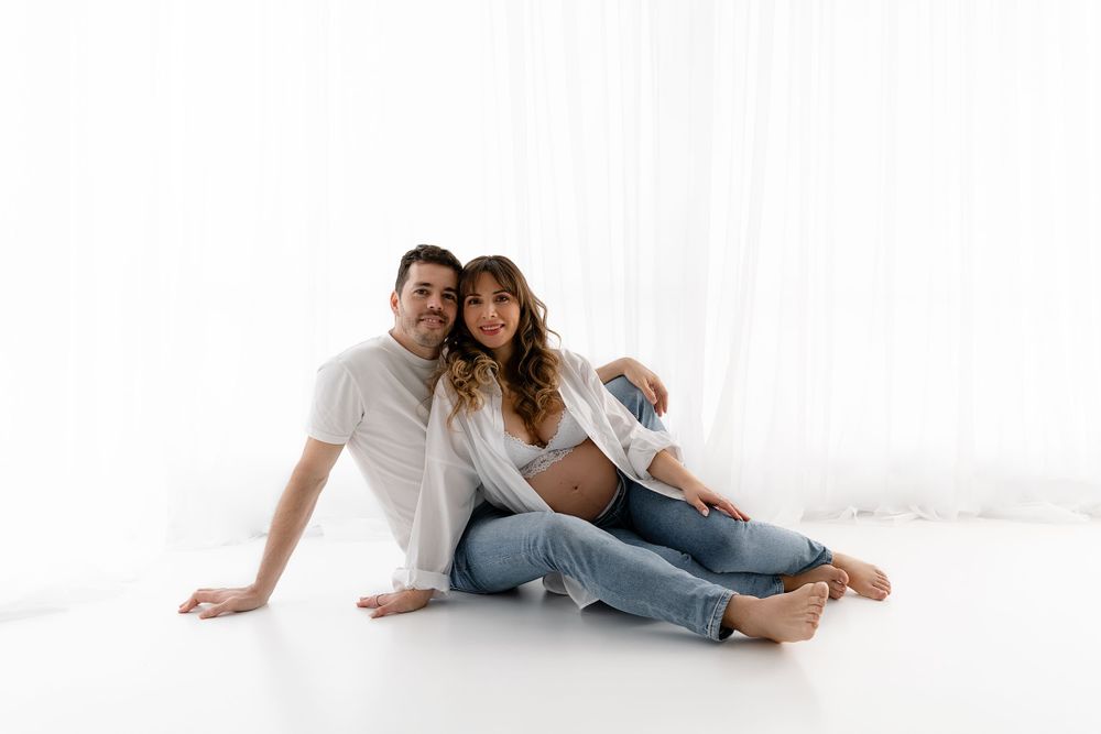 A casual couple in jeans and white shirts sitting together on a white floor.