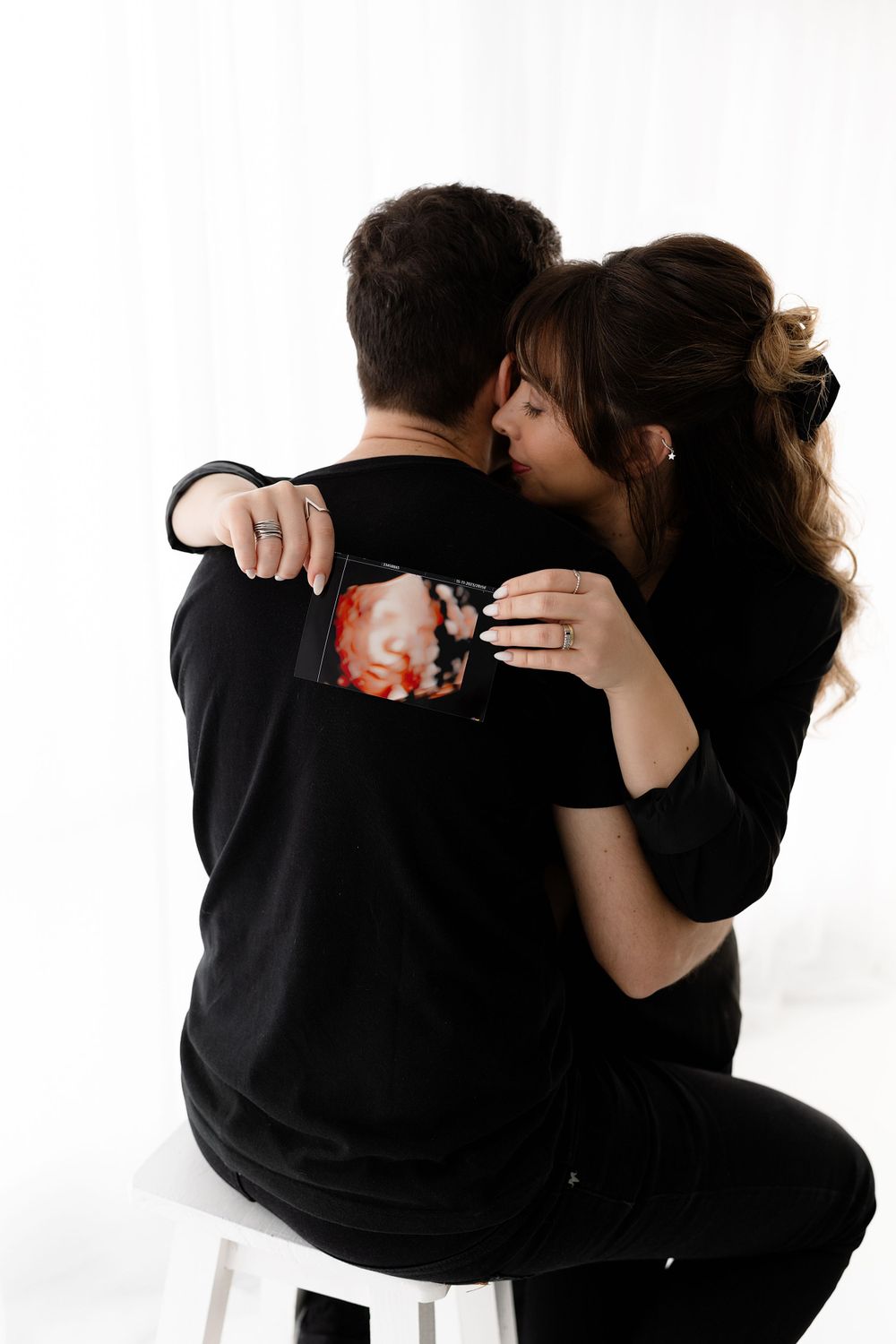 A couple in black clothing embracing while sitting on a white stool.