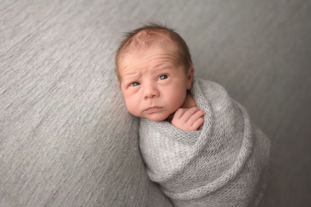 Alert newborn baby wrapped in gray knit swaddle looking at camera with expressive face.