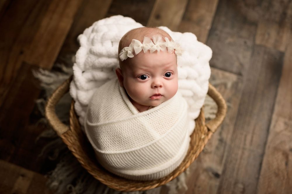 Alert newborn baby girl swaddled in cream wrap with floral headband sitting in wicker basket.