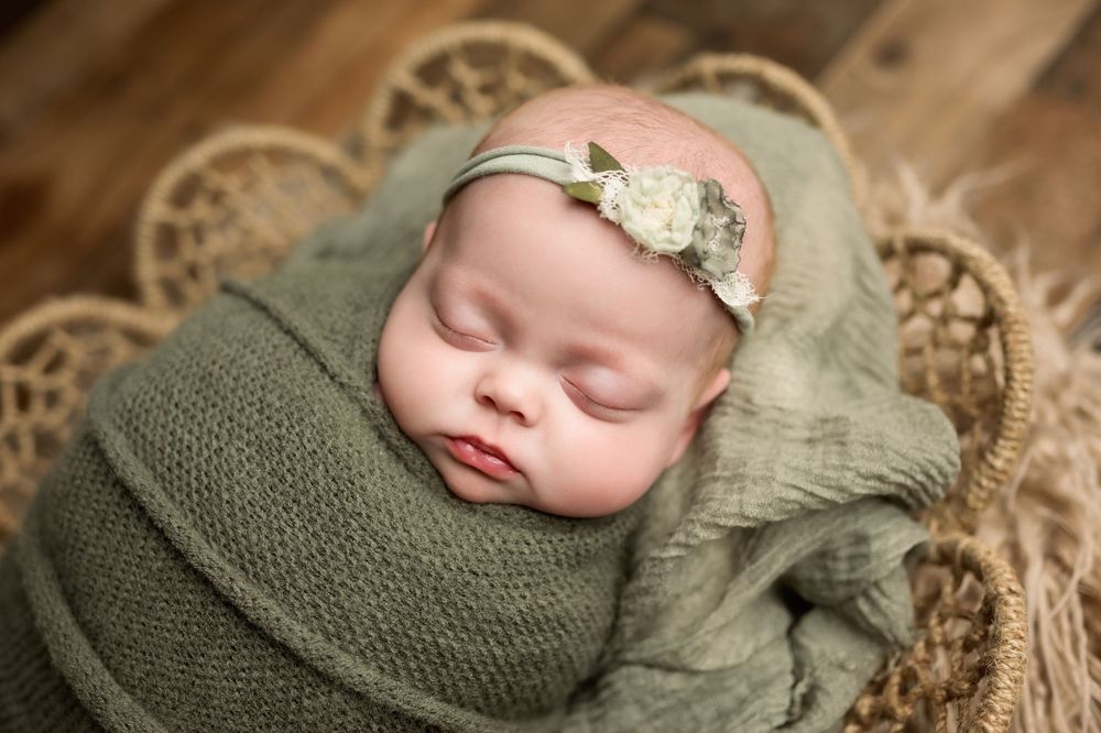 Sleeping newborn baby girl swaddled in sage green wrap with floral headband in wicker basket.