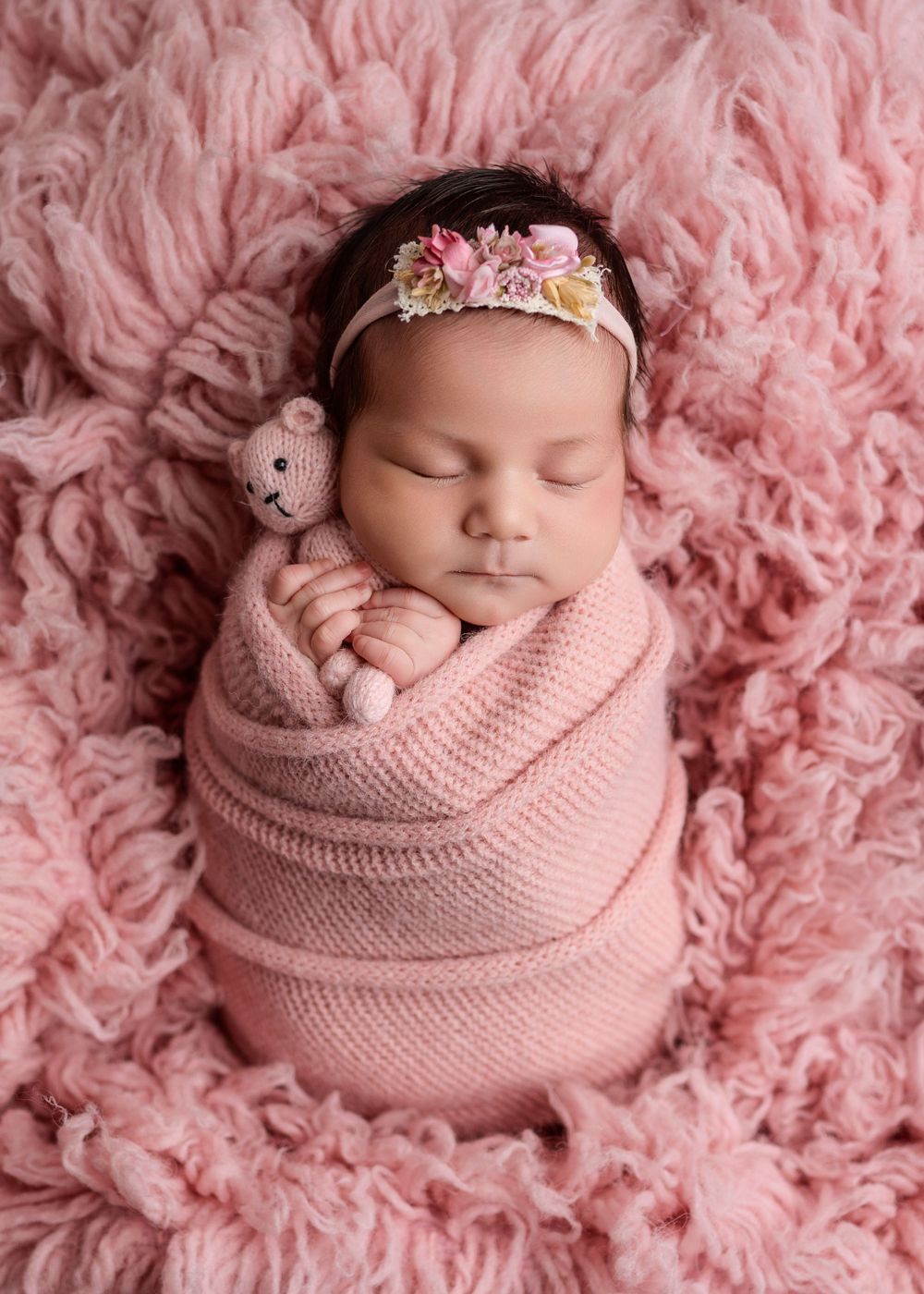 Swaddled newborn baby girl in pink wrap with floral headband holding knit teddy bear on pink fluff.